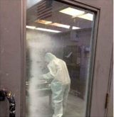 An Ames worker wears coveralls and a full face respirator while applying Conformal coating to a circuit board inside the walk-in paint booth in N211.